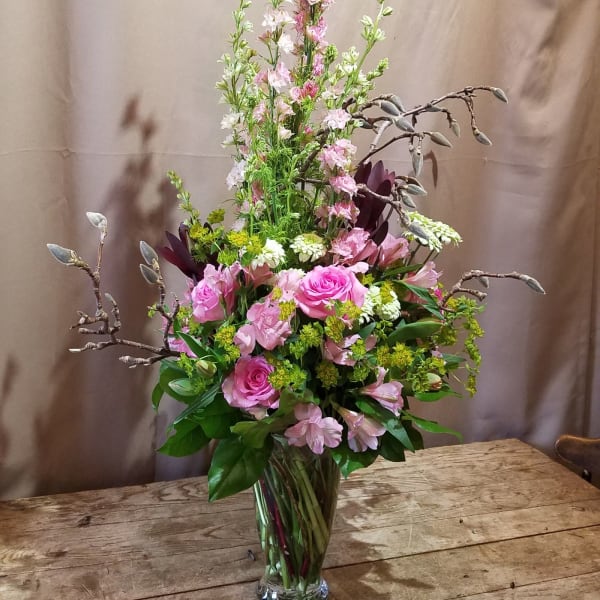 Tall pink and green flower arrangement with roses and delphinium in a clear glass vase on a wooden table.
