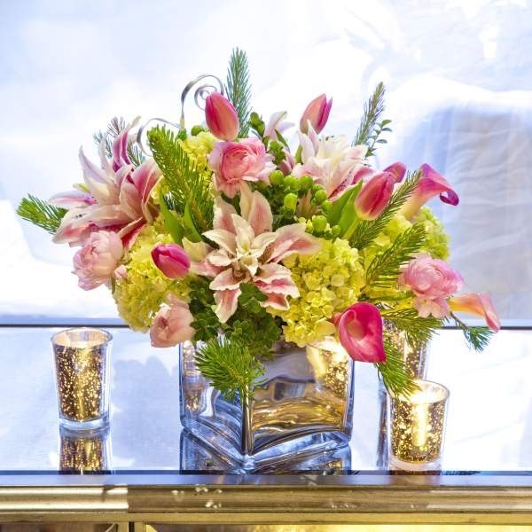 Pink tulips and lilies arranged in a square glass vase with candles