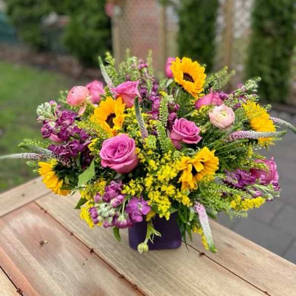 Mixed bouquet of pink roses, yellow sunflowers, and purple flowers in a purple vase