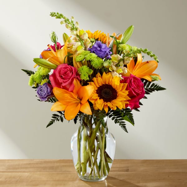 Mixed bouquet of sunflowers, roses, lilies, and purple blooms in a glass vase