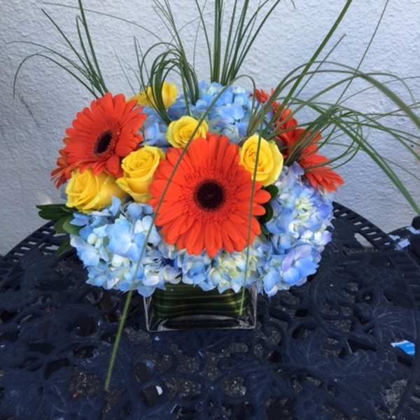 Bright bouquet of orange gerbera daisies, yellow roses, and blue hydrangeas in a glass vase