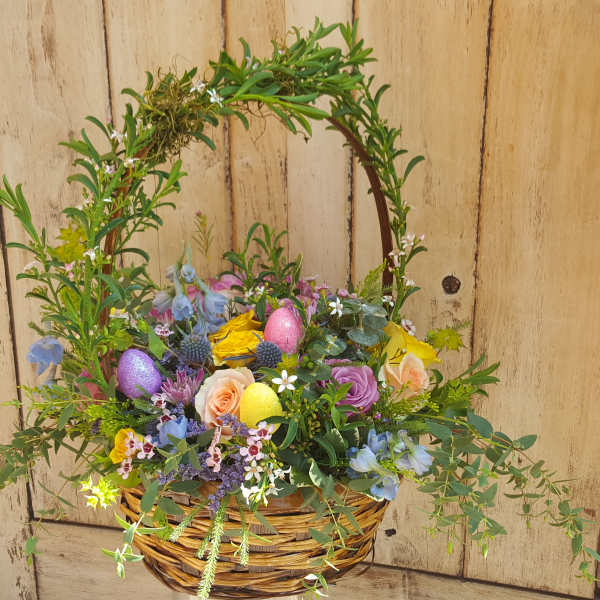 Wicker basket of pastel roses and mixed flowers with a leafy arch and colorful glitter Easter eggs.