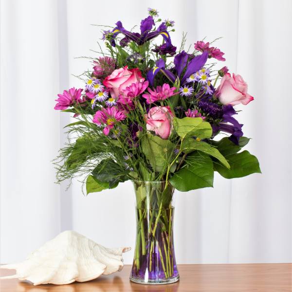 Tall bouquet of pink roses, purple irises, and pink daisies in a clear vase beside a large white seashell.