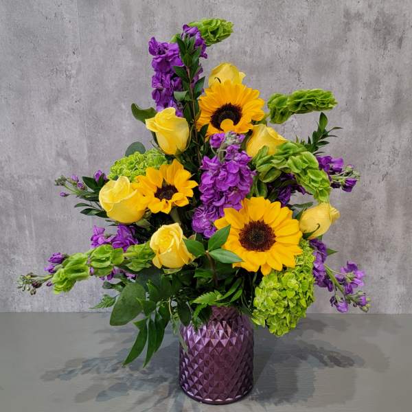 Purple vase holding yellow roses, sunflowers, green hydrangeas, and purple blooms