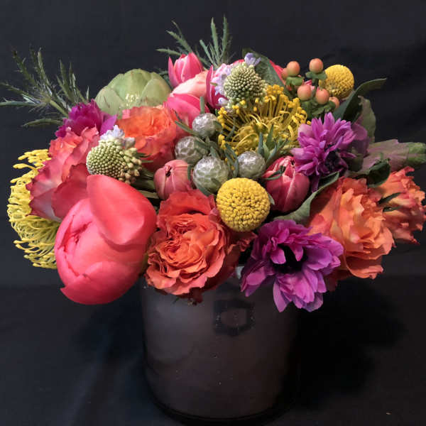 Bright mixed arrangement of pink, orange, yellow, and purple flowers in a smoky glass cylinder vase