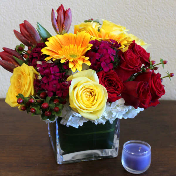 Colorful bouquet of roses and daisies in a square glass vase beside a blue candle