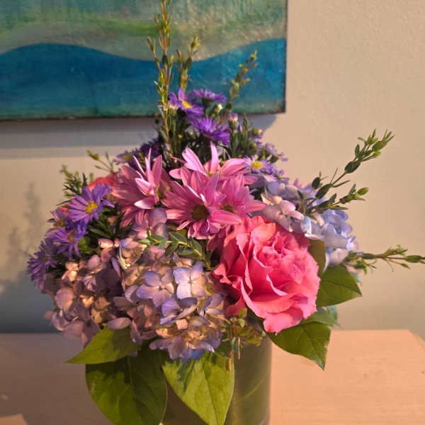 Compact arrangement of pink rose, purple daisies, and lavender hydrangeas in a clear glass cylinder vase