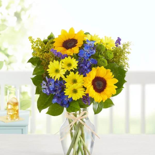 Sunflowers and blue flowers in a clear glass vase with a ribbon