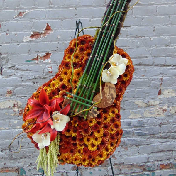 Tall floral arrangement shaped like a guitar with orange mums, red lilies, and white orchids on an easel.