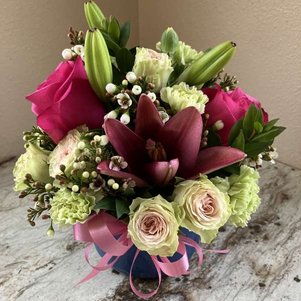 Compact arrangement of pink lilies, roses, and green carnations in a blue vase with a pink ribbon bow