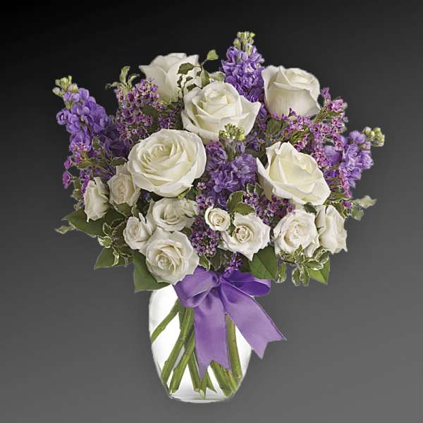 White roses and purple flowers arranged in a glass vase with a purple ribbon.