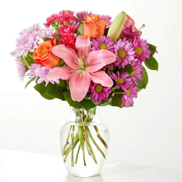 Pink lilies, roses, and purple daisies in a clear glass vase