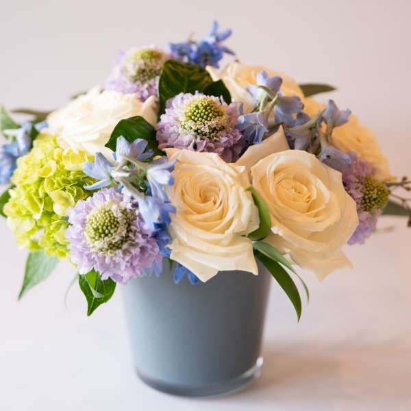 Bouquet of cream roses, blue flowers, and lavender blooms in a gray vase