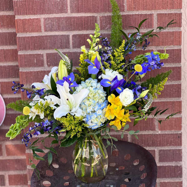 Mixed bouquet of white lilies, blue irises, and yellow flowers in a glass vase.