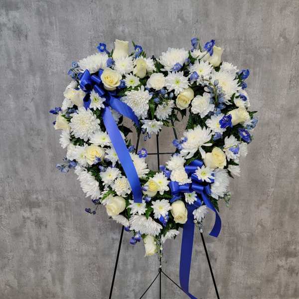 Heart-shaped floral wreath with white flowers, cream roses, and blue ribbons on a stand