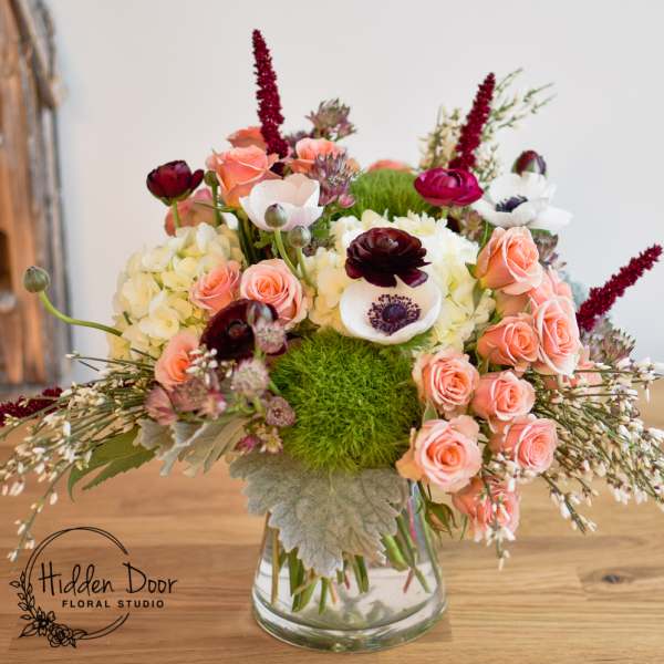 Mixed bouquet of peach roses, white anemones, and burgundy blooms in a glass vase