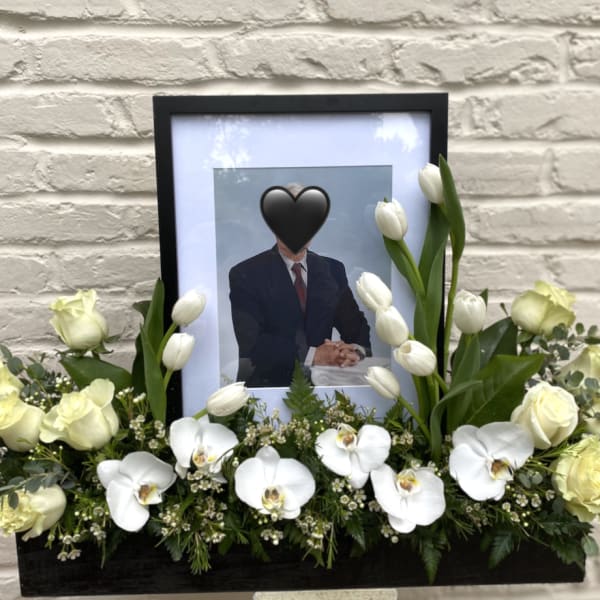 White floral tribute around a framed portrait in a black container.
