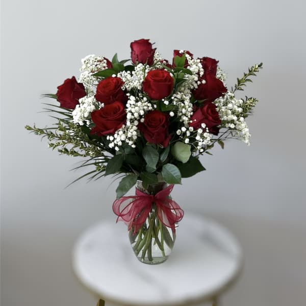 Clear glass vase of red roses with white filler flowers and a red bow.