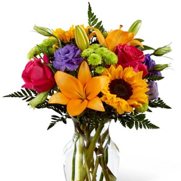 Bright mixed bouquet of lilies, roses, sunflowers, and purple blooms in a glass vase