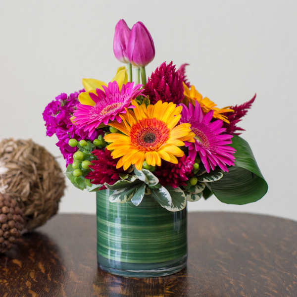 Bright mixed bouquet in a green glass vase with pink, yellow, and magenta flowers.