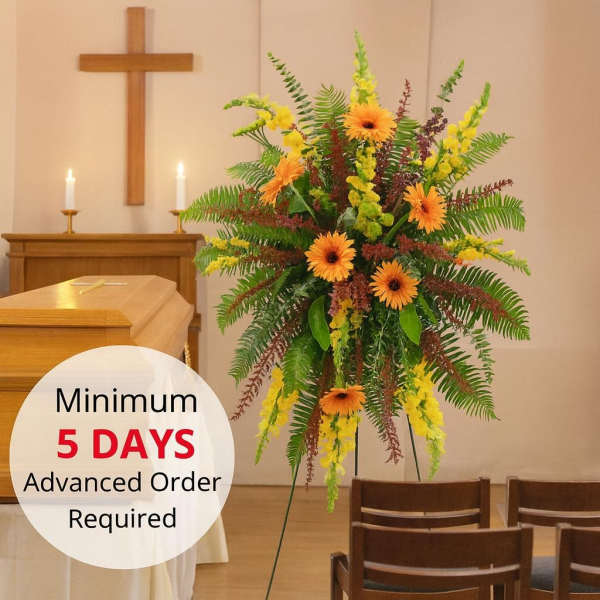 Tall sympathy easel spray with orange gerbera daisies, yellow blooms, and ferns beside a casket.