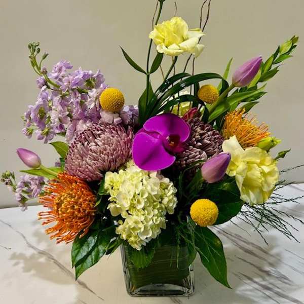 Mixed floral arrangement in a square glass vase with purple, yellow, orange, and pink blooms