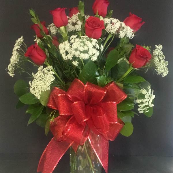 Tall arrangement of red roses and white filler flowers in a clear glass vase with a large red bow.