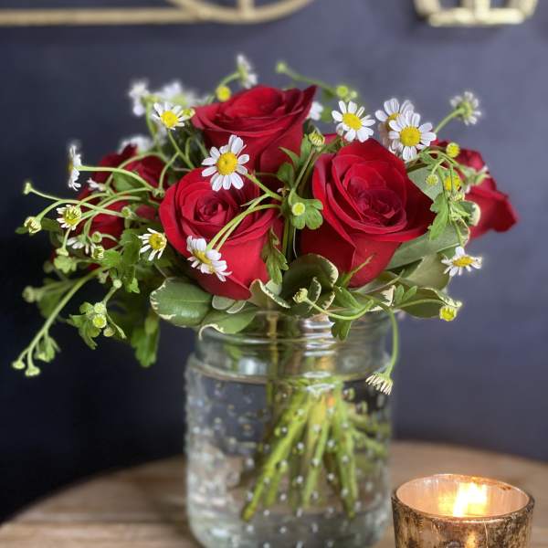 Red roses and white daisies in a glass jar beside a lit candle