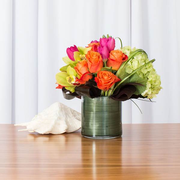 Bright orange and pink flowers with green hydrangeas in a glass cylinder vase wrapped in green leaves.