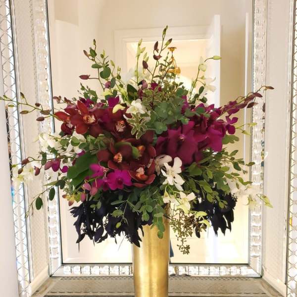 Tall floral arrangement in a gold vase with magenta, burgundy, and white orchids