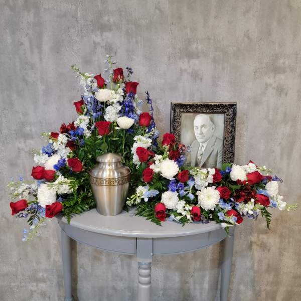 Sympathy floral arrangement with red roses, white blooms, and a memorial urn beside a framed portrait