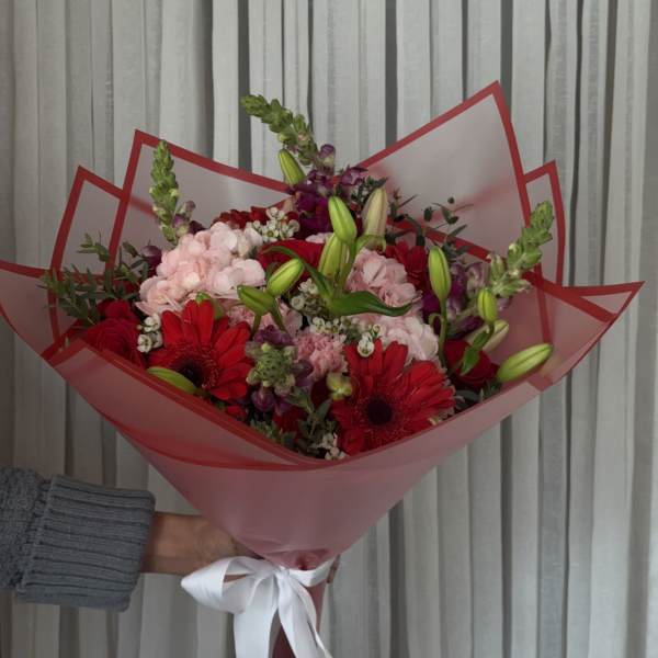 Handheld bouquet of red and pink flowers wrapped in pink paper with a white ribbon