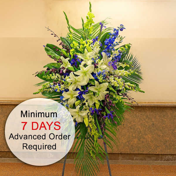 Tall standing spray with white lilies, blue flowers, and bold palm fronds on a black easel.