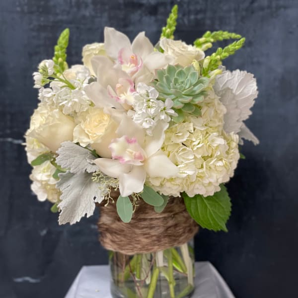 White hydrangeas, roses, orchids, and a succulent in a glass vase wrapped with brown twine