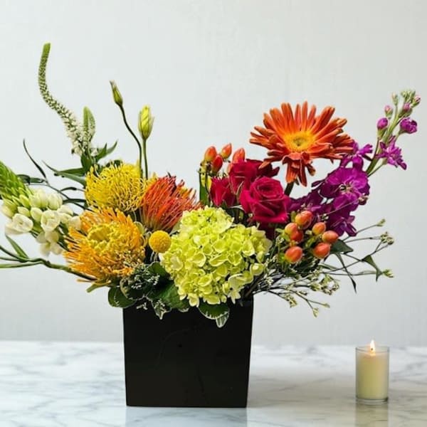 Colorful mixed flower arrangement in a black square vase with a candle beside it