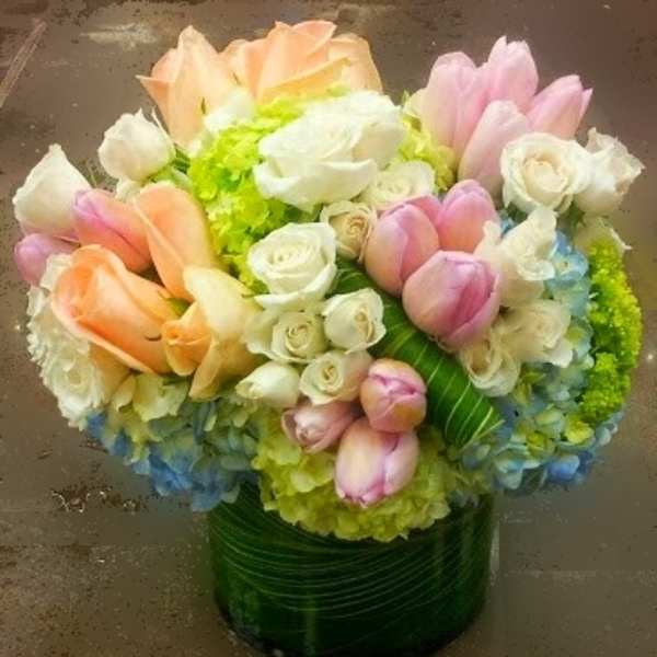 Round arrangement of peach and pink tulips, white roses, and hydrangeas in a leaf-wrapped glass vase