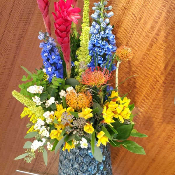 Tall mixed bouquet in a textured glass vase with pink, blue, yellow, and orange flowers