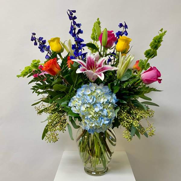 Colorful mixed bouquet in a clear glass vase with hydrangea, roses, lilies, and delphinium.