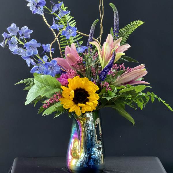Tall mixed arrangement with pink lilies, blue delphinium, and a sunflower in an iridescent vase