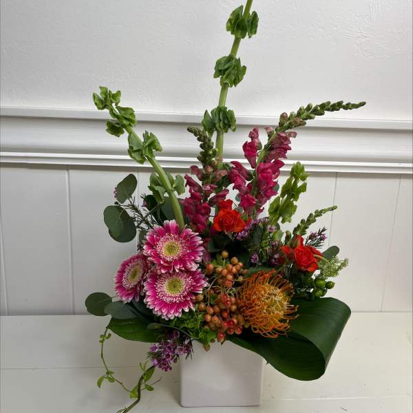 Tall modern arrangement with pink gerberas, snapdragons, orange protea and roses in a white cube vase