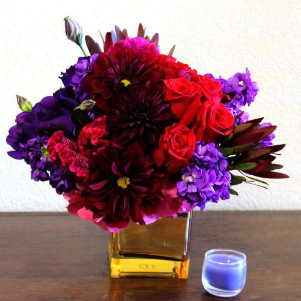 Bouquet of red, purple, and burgundy flowers in a gold vase beside a blue candle