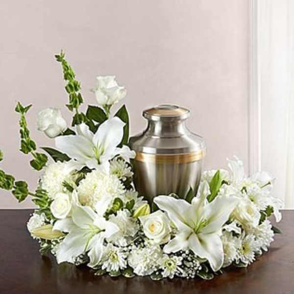 White lilies, roses, and mums arranged in a ring around a silver urn on a dark table.