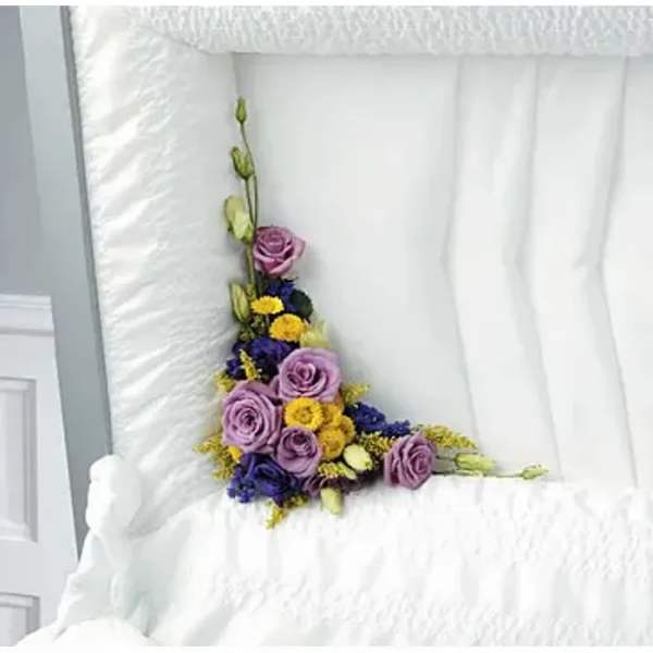 Purple roses and yellow flowers arranged in a white quilted casket interior.