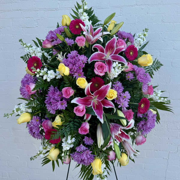 Large standing spray with pink lilies, roses, yellow roses, purple mums, and white accents on an easel.