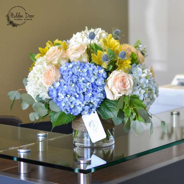 Bouquet of blue hydrangeas, pale roses, and yellow flowers in a glass vase