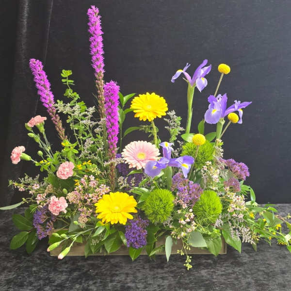 Low rectangular arrangement with yellow gerbera daisies, purple irises, pink carnations, and magenta flower spikes