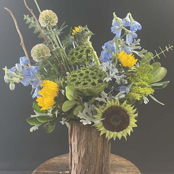 Mixed bouquet with sunflowers, blue flowers, and green textured blooms in a rustic wood vase