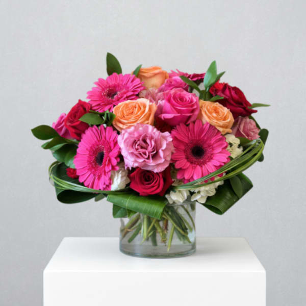 Low round arrangement of pink gerbera daisies and roses with peach accents in a clear glass vase.