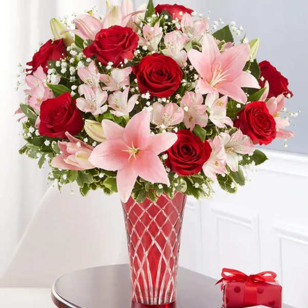 Red roses and pink lilies in a red glass vase beside a small gift box