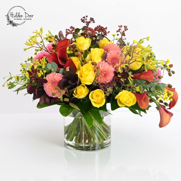 Bouquet of yellow roses, pink blooms, and red calla lilies in a glass vase
